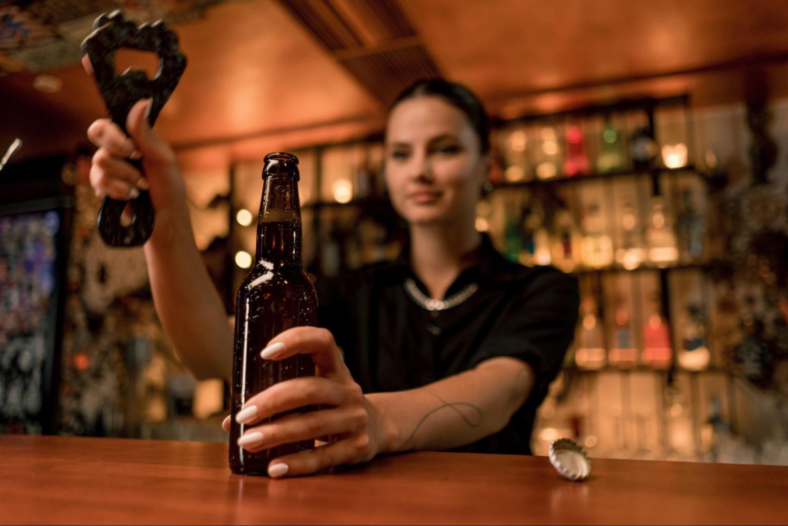 Female bartender at a nightclub bar opening a sealed beer bottle behind the counter