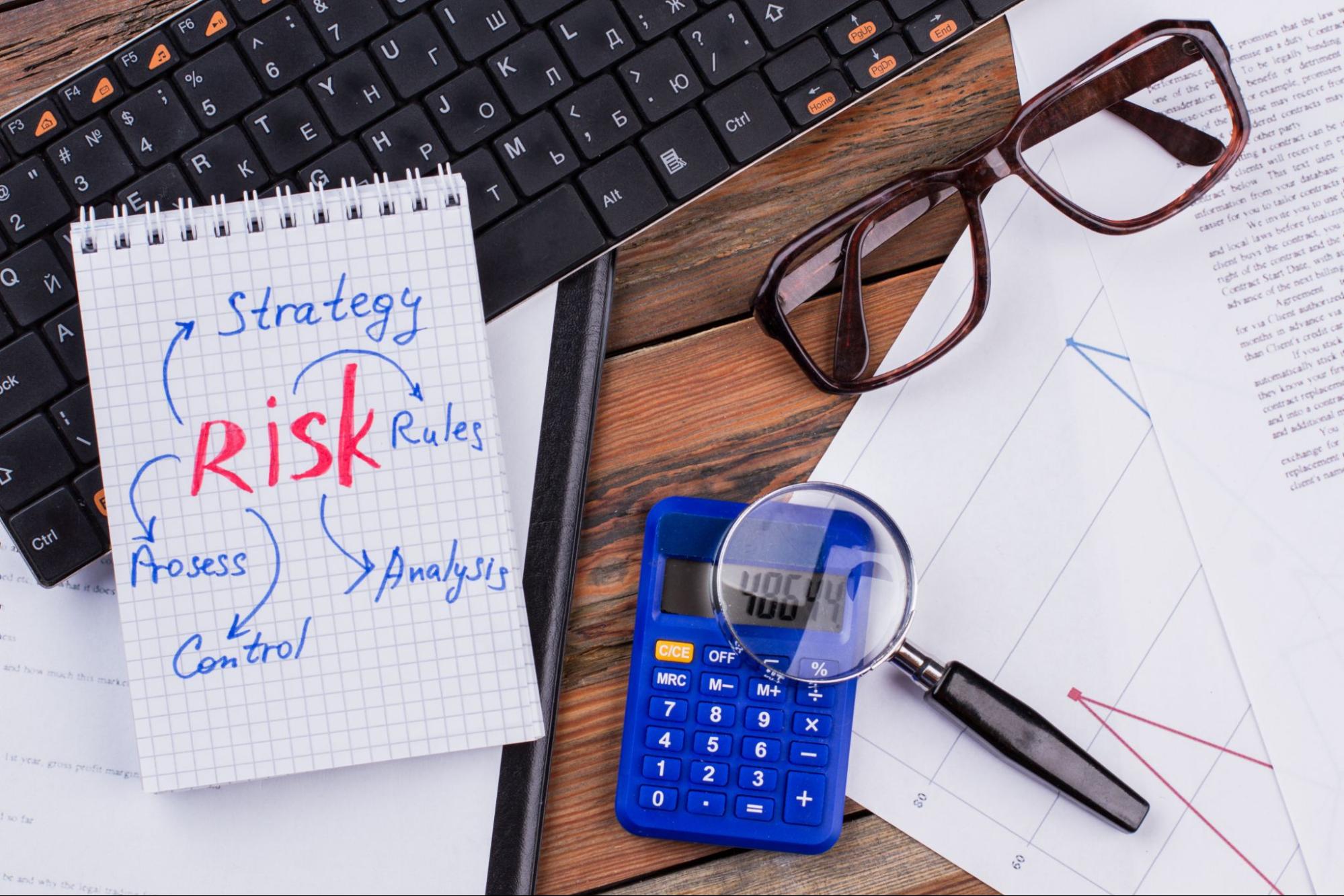 Flat lay of a risk and strategy plan notebook with documents, a magnifier on a blue calculator, and a keyboard arranged on a wooden background