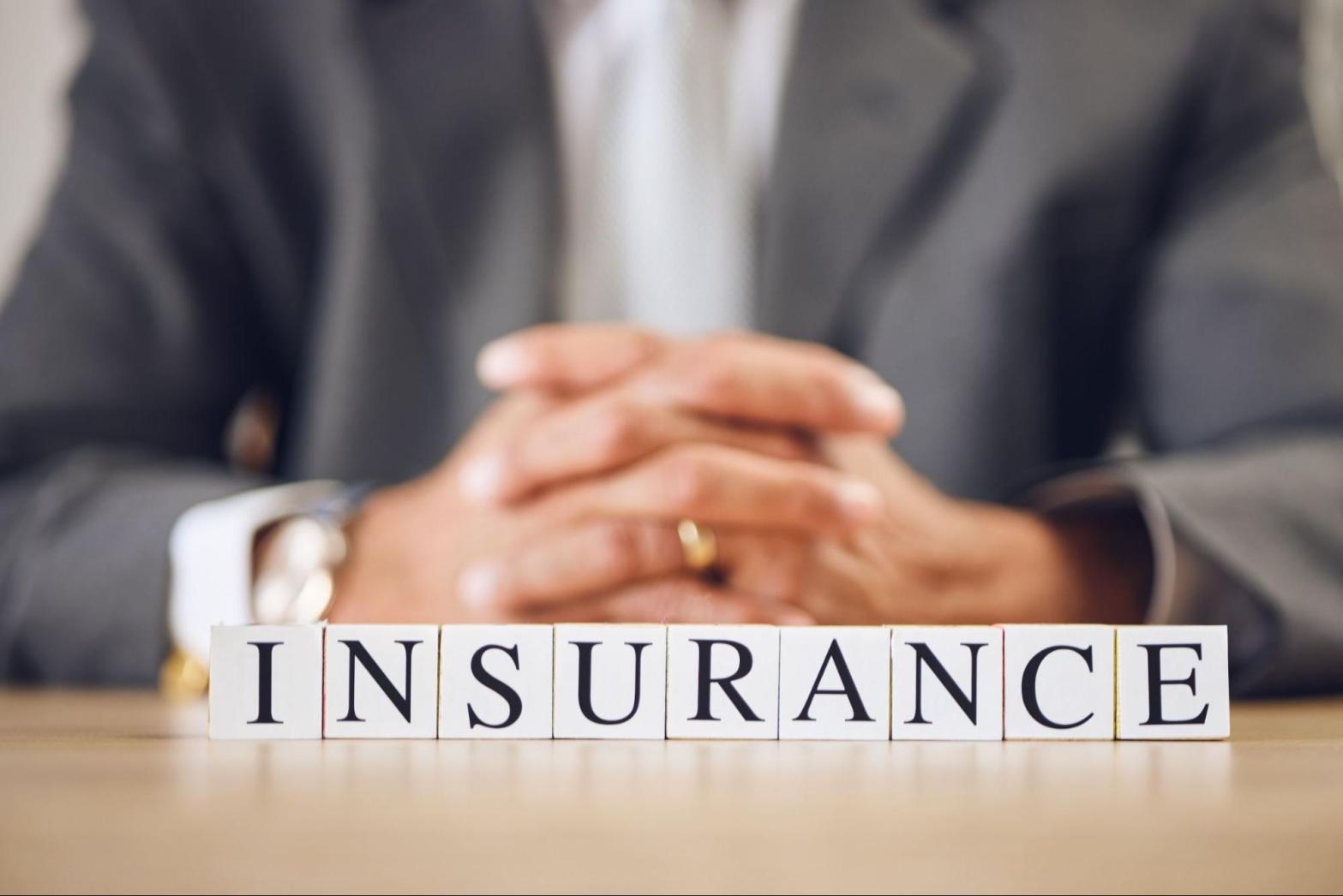 A close-up of a commercial insurance agent’s clasped hands behind the letter blocks spelled “INSURANCE’ on a desk