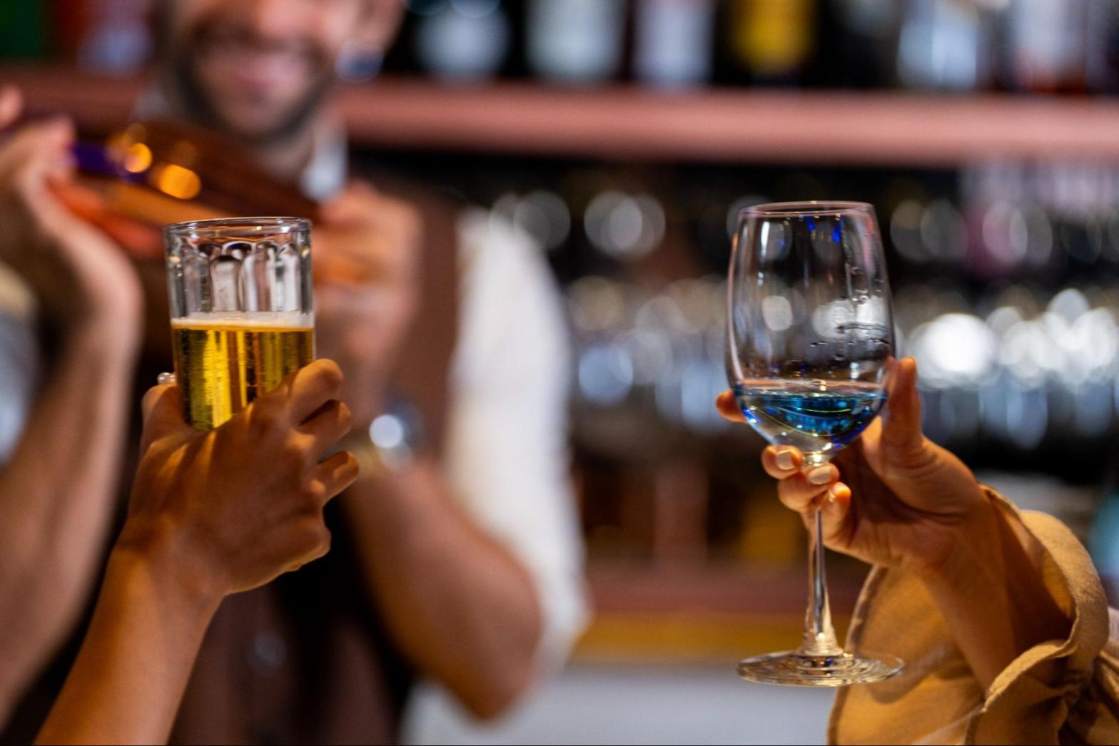 A blurred smiling bartender shakes a cocktail shaker behind the bar as two unseen customers hold alcoholic drinks in front of him