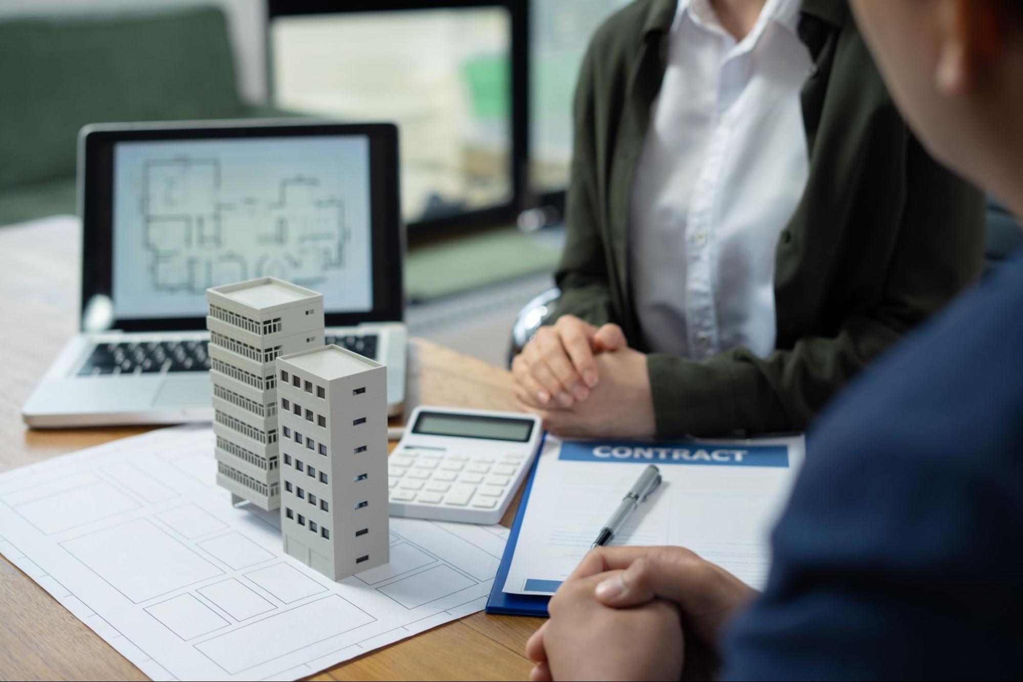 A broker discussing commercial property insurance decisions with a client using building models, a laptop, calculator, and documents